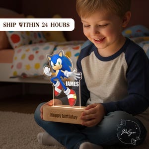 May include: A personalized Sonic the Hedgehog night light. The blue and white character is mounted on a wooden base with the text "Happy birthday" and a name. The light is glowing, and the child is smiling.