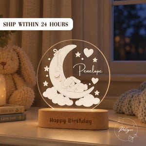 May include: A circular acrylic night light featuring an engraved sleeping bunny on a crescent moon, stars, and hearts. The name Penelope is etched on the surface. The light sits on an oval wooden base with the text Happy Birthday engraved on it.