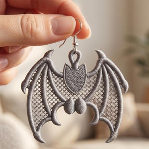 May include: A silver-colored bat-shaped earring with intricate embroidered details. The earring features a detailed wing design and a shield-like shape at the top. The earring is held up by a silver hook.