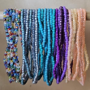 May include: A collection of beaded necklaces in various colors, including blue, purple, and peach, hanging from a wooden dowel. The beads are small and strung together, creating a textured appearance. The necklaces are displayed against a neutral background.