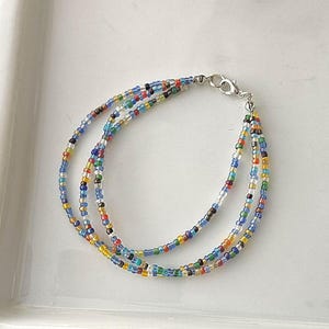 May include: A multi-strand beaded bracelet with a silver clasp. The bracelet features small, colorful beads in shades of blue, yellow, red, green, and black. The bracelet is displayed on a white surface.