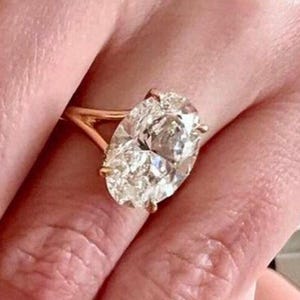 May include: A gold engagement ring featuring a large, oval-cut diamond. The ring's band is a delicate gold, and the diamond is set with prongs. The ring is shown on a person's finger.