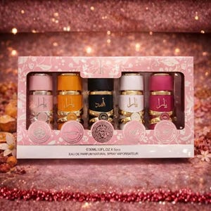 Peut inclure: Un coffret rose contenant cinq flacons de parfum de différentes couleurs : rose, orange, noir, blanc et magenta. Chaque flacon a des accents dorés et une écriture arabe. La boîte a un motif floral rose et le texte "EAU DE PARFUM NATURAL SPRAY VAPORISATEUR".