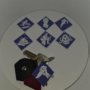 Può includere: Un mazzo di chiavi con un portachiavi in pelle nera, un'etichetta rossa e un'etichetta blu con un pugno bianco. Le chiavi sono su una superficie circolare bianca con sei grafiche a forma di diamante blu.
