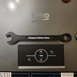 May include: A black wrench-shaped magnet featuring the white text Happy Fathers Day centred on the handle. The tool-themed magnet is positioned horizontally on a metallic grey surface, flanked by two circular planet-themed magnets.