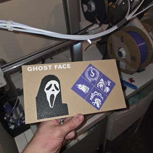 May include: A rectangular tan card featuring the white text GHOST FACE, a black and white Ghost Face mask graphic, and a purple square containing four white icons depicting a hook, a bird, a screaming face, and a hand reaching out.
