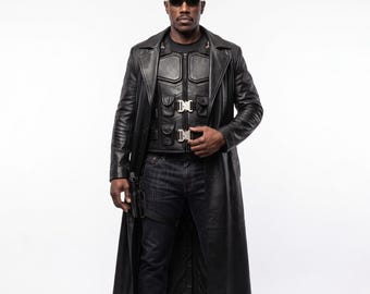 May include: A person wearing a long black leather trench coat over a black tactical vest with silver buckles and pockets. The outfit includes dark blue denim jeans and dark sunglasses. A black holster is attached to the right side of the waist.