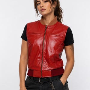 May include: A sleeveless red leather vest featuring a brown ribbed collar, a central metal zipper closure, and a dark red ribbed hem. The vest is worn over a plain black short-sleeved t-shirt, paired with black trousers and a silver chain accessory.