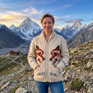 May include: A cream-colored zip-up sweater with a fox design in red and brown on the front. The sweater has pockets and blue accents on the sleeves and pockets. The background features a mountain range and a blue sky.