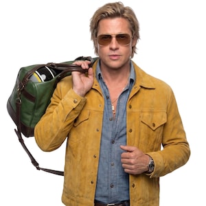 Könnte beinhalten: Eine Person trägt eine senfgelbe Wildlederjacke über einem blauen Hemd, gold getönte Fliegerbrille und eine silberne Armbanduhr. Über der Schulter wird eine grüne und braune Ledertasche getragen, in der sich ein Helm mit gelben Streifen befindet.