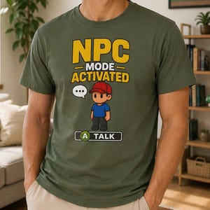 May include: Olive green t-shirt with the text "NPC MODE ACTIVATED" in yellow. A cartoon character wearing a red cap and blue shirt is featured below the text, with a speech bubble and the word "TALK".