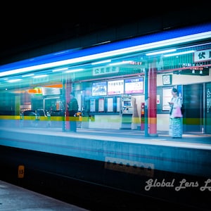Könnte beinhalten: Nachtaufnahme eines japanischen Bahnhofs mit einem verschwommenen Zug in Bewegung. Der Bahnhof hat blaue und grüne Neonlichter. Eine Person steht auf dem Bahnsteig und schaut auf ihr Telefon. Langzeitbelichtungseffekt.