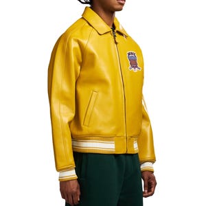 May include: A yellow leather jacket with a zip-up front, a collar, and a patch on the chest. The jacket has white stripes on the cuffs and hem. The patch features a red, white, and blue design with the word "AIRWAY".