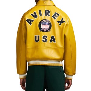 May include: Mustard yellow leather jacket with the text "AVIREX" and "USA" on the back. The jacket features a patch with the American flag and the words "ALL AMERICANS ESTABLISHED 1975". The jacket has white and black striped cuffs.