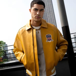 May include: Mustard yellow leather bomber jacket with a white and black striped hem. The jacket features a patch with a red, white, and blue design and the word "AIREX". The jacket has a classic collar and a zipper closure.