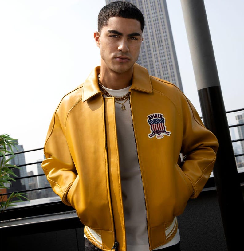 May include: Mustard yellow leather bomber jacket with a white and black striped hem. The jacket features a patch with a red, white, and blue design and the word "AIREX". The jacket has a classic collar and a zipper closure.