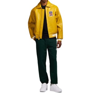 May include: A yellow leather jacket with a zip closure and a small patch on the upper left chest. The jacket has white and cream-colored striped cuffs and a matching hem. The person is wearing a black shirt and dark green sweatpants.