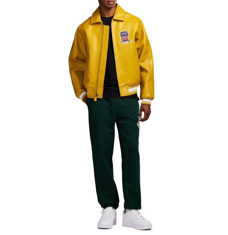 May include: A yellow leather jacket with a zip closure and a small patch on the upper left chest. The jacket has white and cream-colored striped cuffs and a matching hem. The person is wearing a black shirt and dark green sweatpants.