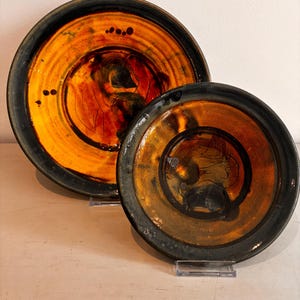 Peut inclure: Deux assiettes rondes en céramique avec un vernis brillant, présentant un centre orange et jaune vif qui se fond dans un bord noir anthracite. La surface est ornée de motifs linéaires sombres et de petits points circulaires sur l'émail.