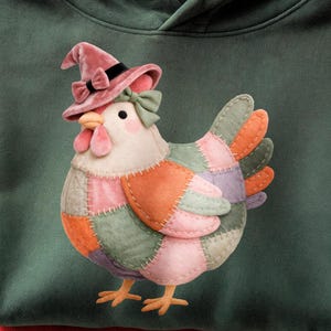 May include: A dark green hooded sweatshirt featuring a central graphic of a chicken wearing a pink witch hat with a black band and bow. The chicken has a patchwork-style body in shades of pink, orange, purple, and sage green with visible stitching.