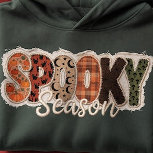 May include: A dark olive green hooded sweatshirt featuring a large, raw-edge fabric appliqué across the chest. The appliqué spells out the word SPOOKY in patterned letters with pumpkins, black cats, moons, plaid, webs, and mushrooms, with Season below.