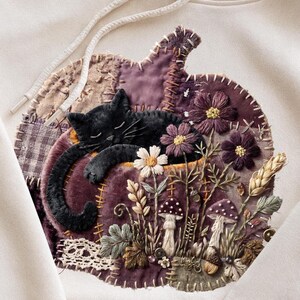 May include: A cream-colored hoodie featuring a large, circular fabric appliqué shaped like a pumpkin. The design includes a black cat sleeping on the pumpkin, surrounded by embroidered flowers, mushrooms, wheat stalks, and lace trim details.