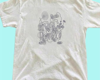 Op de afbeelding: Een wit T-shirt met korte mouwen en een centrale grafische print in zwarte inkt. Het ontwerp bestaat uit ingewikkelde, wervelende lijntekeningen die lijken op paddenstoelen of abstracte organische vormen, met de tekst Mac DeMarco in het midden.