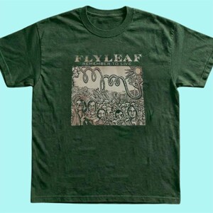 Puede incluir: Una camiseta de manga corta de color verde oscuro con un estampado gr&aacute;fico en el pecho. El estampado muestra el texto "FLYLEAF" y "REMEMBER TO LIVE" sobre una ilustraci&oacute;n de una serpiente enroscada y una multitud de personas.