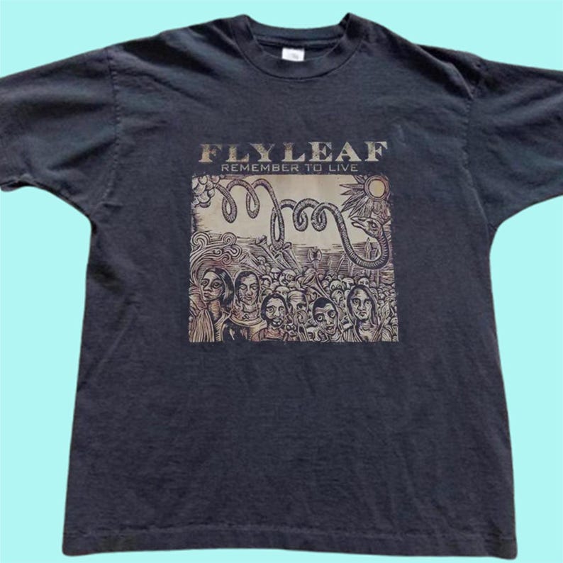Puede incluir: Una camiseta negra de manga corta con un estampado gr&aacute;fico que incluye el texto "FLYLEAF" y "REMEMBER TO LIVE" sobre una ilustraci&oacute;n de una multitud, una serpiente enroscada y un sol, todo en tonos de tinta marr&oacute;n y beige.