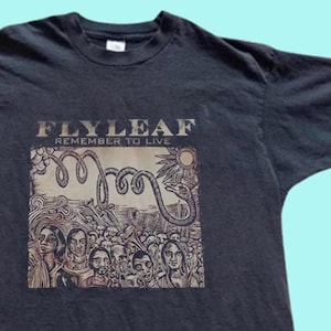 Puede incluir: Una camiseta negra de manga corta con un estampado gr&aacute;fico en el pecho. El estampado muestra el texto "FLYLEAF" y "REMEMBER TO LIVE" sobre una ilustraci&oacute;n de una multitud de personas, una gran serpiente enroscada y un sol en color beige.