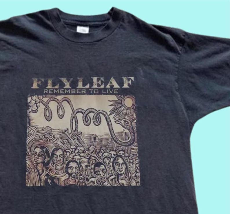 Puede incluir: Una camiseta negra de manga corta con un estampado gr&aacute;fico en el pecho. El estampado muestra el texto "FLYLEAF" y "REMEMBER TO LIVE" sobre una ilustraci&oacute;n de una multitud de personas, una gran serpiente enroscada y un sol en color beige.