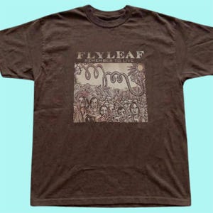 Puede incluir: Una camiseta de manga corta de color marr&oacute;n oscuro con un estampado gr&aacute;fico en el pecho. El estampado muestra el texto FLYLEAF REMEMBER TO LIVE sobre una ilustraci&oacute;n de una multitud de personas, una serpiente enroscada y un sol.