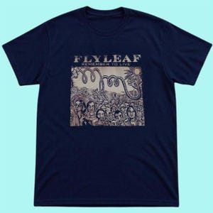 Puede incluir: Una camiseta azul marino de manga corta con un estampado gr&aacute;fico en el pecho. El estampado muestra el texto "FLY LEAF" y "REMEMBER TO LIVE" sobre una ilustraci&oacute;n de una serpiente enroscada, un sol y una multitud de personas.