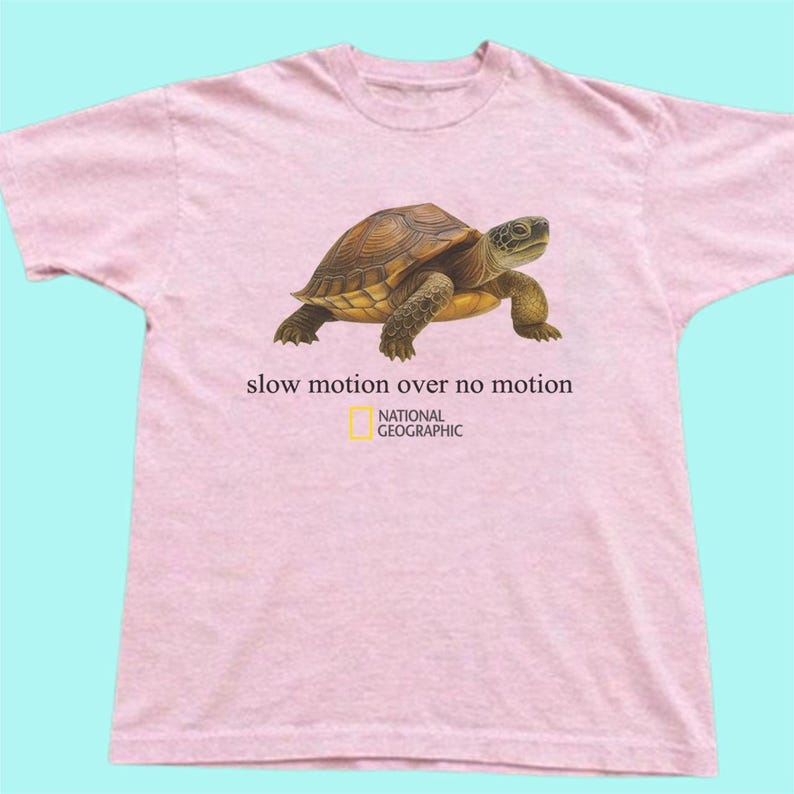 May include: A light pink short-sleeved t-shirt featuring a graphic of a brown turtle. Below the turtle, the text reads "slow motion over no motion" followed by the National Geographic logo, which includes a yellow rectangle and black text.