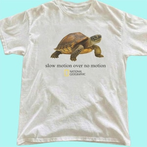 Puede incluir: Una camiseta blanca de manga corta con un gráfico de una tortuga marrón en el centro. Debajo de la tortuga, el texto dice "slow motion over no motion" seguido del logotipo y el nombre de National Geographic en letras negras.