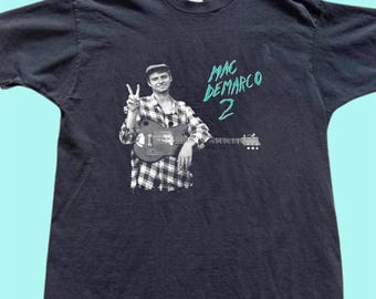 Op de afbeelding: Zwart t-shirt met een zwart-wit afbeelding van een persoon met een gitaar die het vredesteken maakt. De tekst "MAC DEMARCO 2" is in turquoise op de rechterkant van het shirt gedrukt.