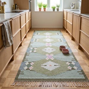 May include: A rectangular, patterned rug with a geometric design in shades of green, blue, and white. The rug has a fringe on the ends and is placed in a kitchen setting. Two pink pet bowls are on the rug.