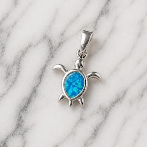 Op de afbeelding: Een zilverkleurige schildpendant met een helderblauwe kern. De hanger heeft een gedetailleerd schildpadontwerp met vinnen en een kop. De blauwe kern heeft een gespikkelde uitstraling. De hanger is ontworpen om als ketting te worden gedragen.