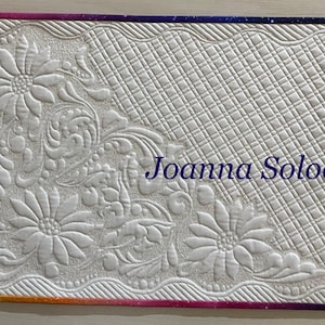 Può includere: Tessuto trapuntato bianco con disegni floreali e volute. Il bordo presenta una sfumatura di colori, dal giallo al viola. Il testo "Joanna Soloch" è visibile sul tessuto.