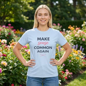 Peut inclure: Un t-shirt bleu clair avec le texte "MAKE sense COMMON AGAIN" en bleu marine et rose. Le t-shirt présente un petit motif de nœud. La personne se tient dans un jardin avec des fleurs colorées.