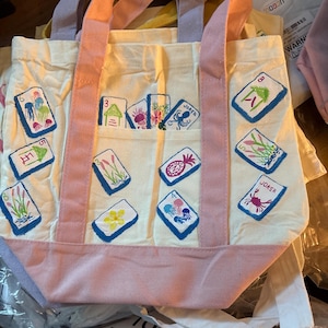 May include: A cream-colored canvas tote bag featuring pink handles and a pink base. The bag is decorated with various embroidered mahjong tile designs, including jellyfish, flowers, and joker tiles, scattered across the front and on a pocket.