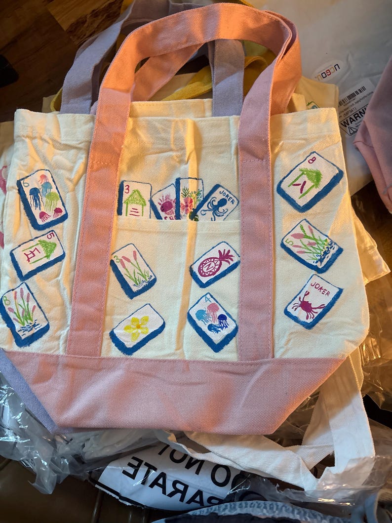 May include: A cream-colored canvas tote bag featuring pink handles and a pink base. The bag is decorated with various embroidered mahjong tile designs, including jellyfish, flowers, and joker tiles, scattered across the front and on a pocket.