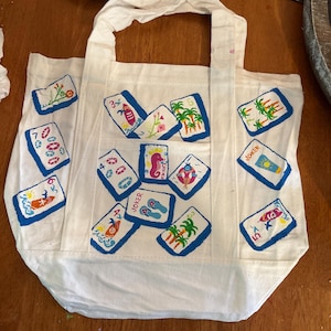May include: A white canvas tote bag featuring a hand-painted pattern of various mahjong tiles. The tiles display colorful illustrations including palm trees, surfboards, seahorses, flip-flops, and life preservers, with some tiles labeled as Joker.