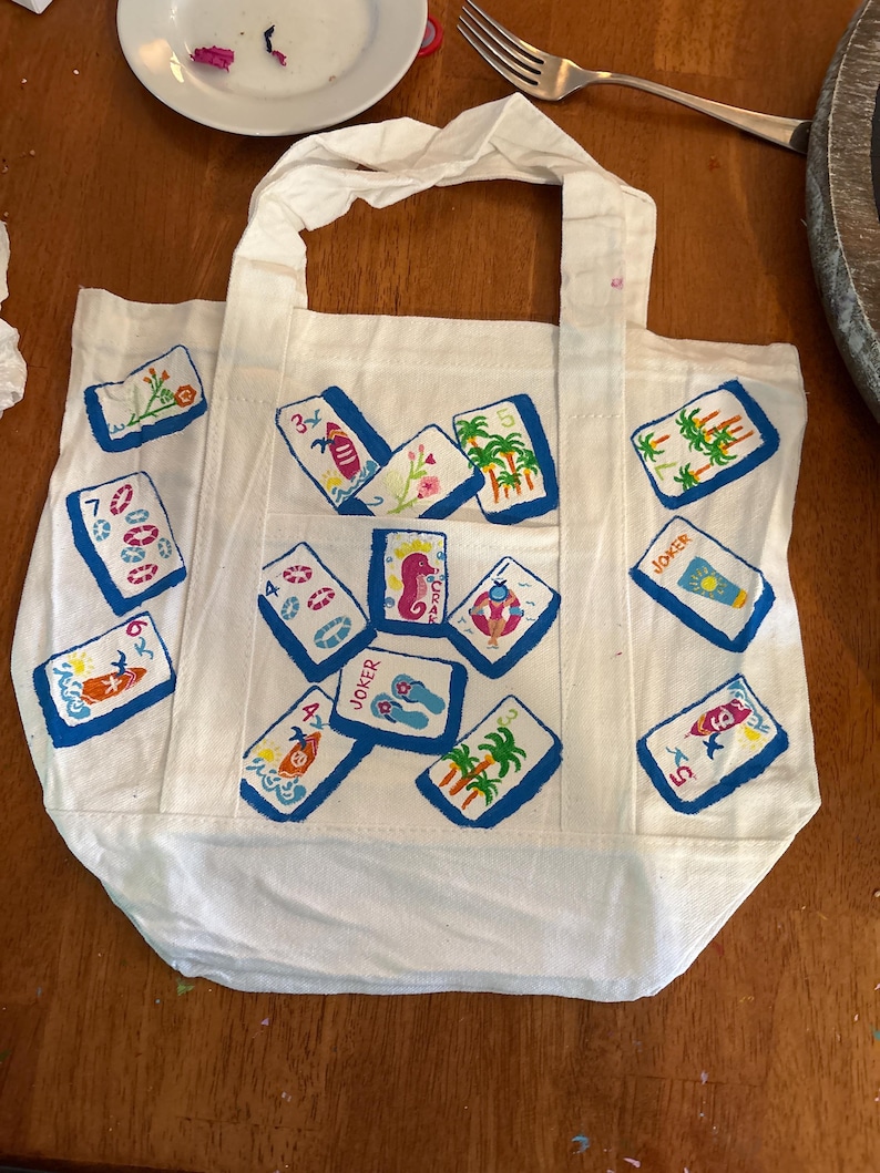 May include: A white canvas tote bag featuring a hand-painted pattern of various mahjong tiles. The tiles display colorful illustrations including palm trees, surfboards, seahorses, flip-flops, and life preservers, with some tiles labeled as Joker.