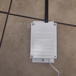 May include: A white rectangular electronic device with a black antenna extending from the top. The front panel features three raised rectangular sections with the embossed text Meshcore, Emergency, and Messaging. A white cable extends from the bottom.