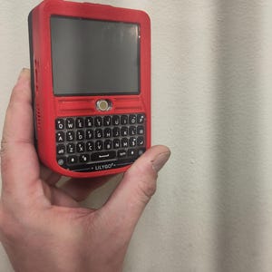 Op de afbeelding: Een rood handheld apparaat met een zwart scherm en een volledig QWERTY-toetsenbord. Het apparaat heeft een zwarte antenne en de merknaam "LILYGO" staat onderaan gedrukt. Het apparaat wordt in een hand gehouden.