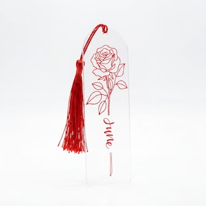 May include: A clear, rectangular acrylic bookmark with rounded top edges, featuring a red line-art illustration of a rose and the word "June" written in a cursive script. A red fabric tassel is attached to the top hole of the bookmark.