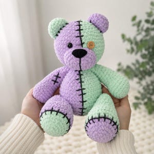 May include: A plush, crocheted teddy bear featuring a split-color design in pastel purple and mint green. The bear has a black stitched seam down the center, a black nose, one black button eye, and one brown wooden button eye on its face.