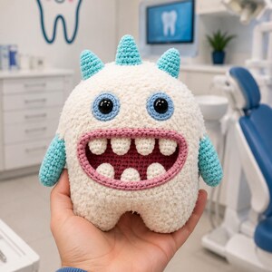 May include: A white crochet monster plush toy with three light blue horns, two black plastic eyes, and a wide open mouth featuring a pink interior and four white teeth on the top and bottom rows. The creature has light blue arms.