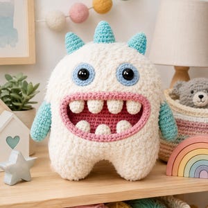 May include: A plush crochet monster toy with a fuzzy white body, light blue horns and arms, and large black eyes. It features a wide, open mouth with a pink interior and several white teeth. The creature has a rounded, soft-textured appearance.